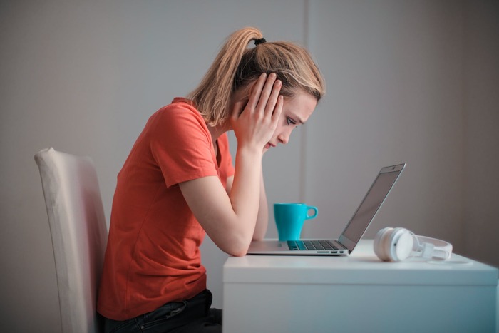 woman in orange shirt sitting in front of a laptop holding her face with two hands looking concerned