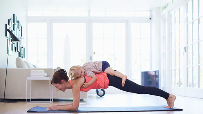 young mom exercising with a toddler on her back on a yoga mat in a living room 