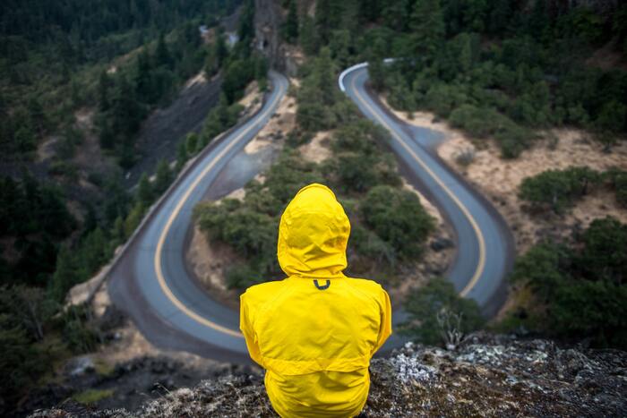 person in a yellow raincoat sitting at a top of a rock watching a road turning in the mountains
