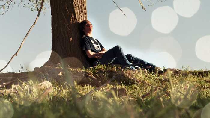 young man sitting against a tree trunk outside enjoying the sunny day