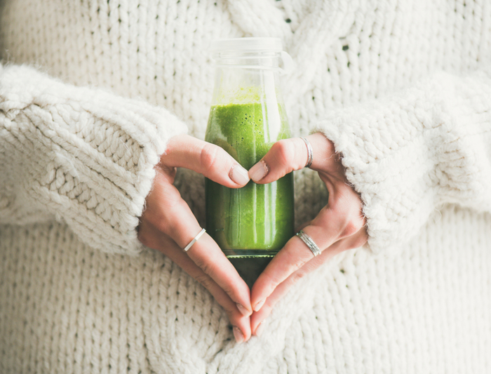 woman dressed in white sweather holding a small bottle of green smoothie in her hands in a heart shape