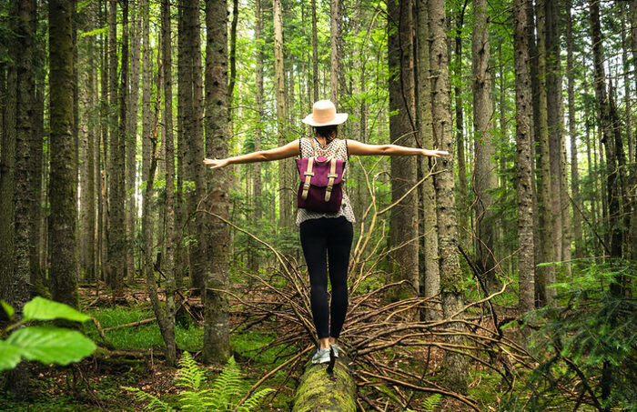 woman outdoors in the forest walking on a fallen tree trunk with her hat and backpack on