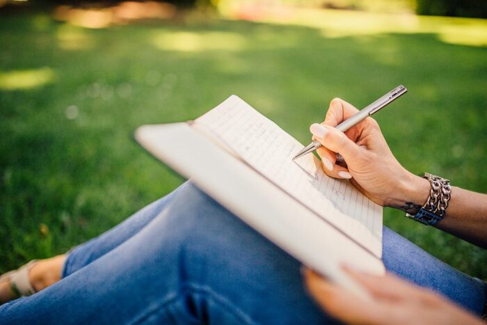 woman dressed in jeans with a metal bracelet on sitting on a green grass field writing in her diary 