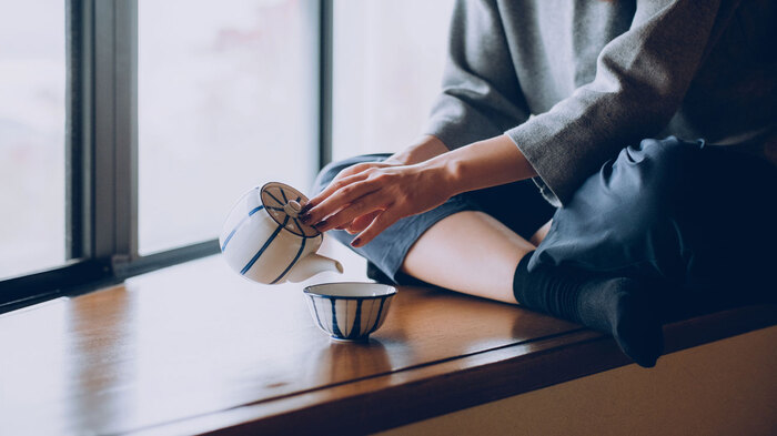 woman in dark clothes sitting on a window sill pouring tea from a white and blue teapot into a small cup