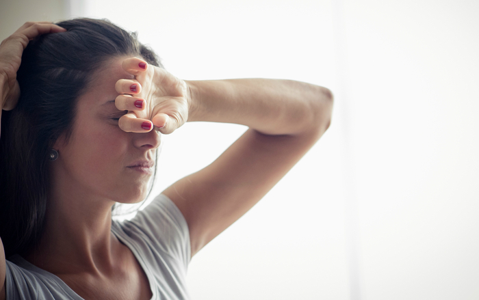 girl having a headache with her hand on her forehand holding her dark hair up and standing against a window