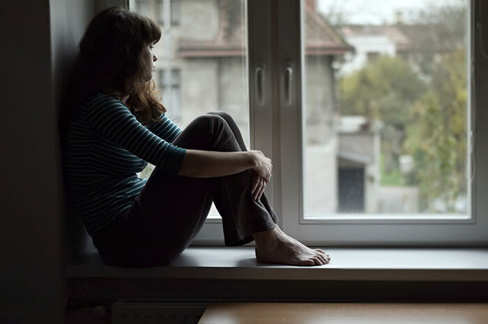 lonely girl sitting on a window sill looking outside with a sad face 