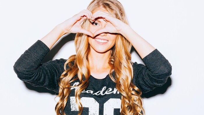 blond woman in a black shirt smiling at the camera making a heart symbol with her hands