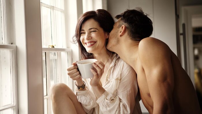 couple morning routine woman sitting at a window smiling holding a white mug and a man kissing her