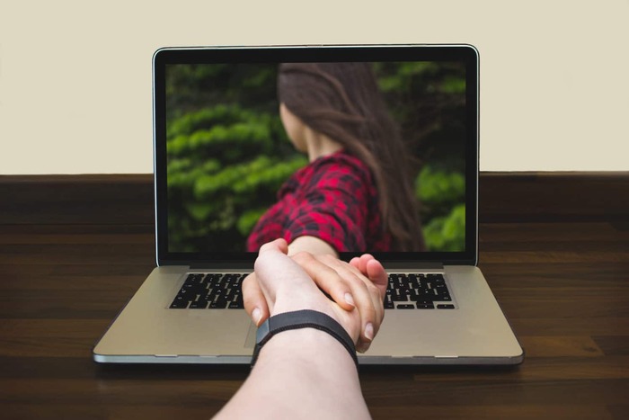 man and woman holding hands through a laptop on a dark wooden surface