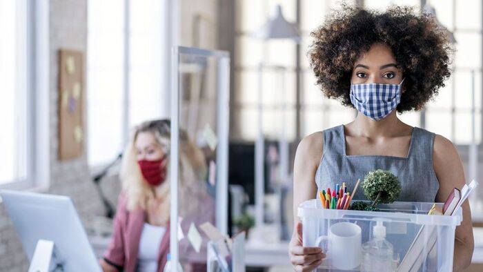 quiting a job woman with curly hair walking in an office environment with a transparent box in her hands and a blue and white mask