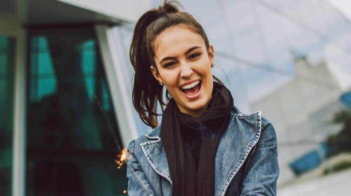 woman with dark hair jeans jacket and black scarf smiling at the camera with glass windows in the background