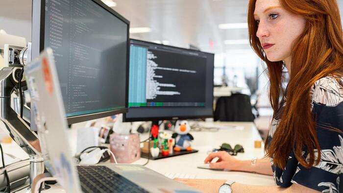 woman with red hair working on a desk with two large monitors in front of her