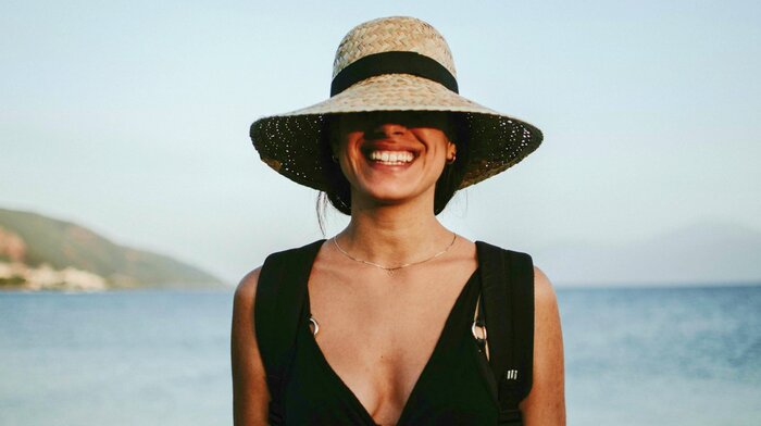 woman in a black swim suit and a sun hat smiling with sea in the background
