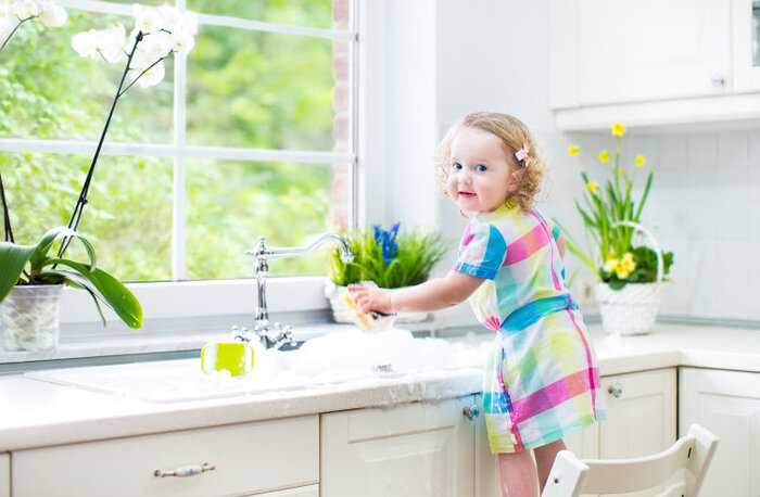 child with colorful dress in kitchen cleaning the counterop in front of a large window with spring flowers around