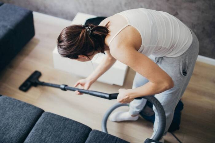 woman in pants and top with her hair pulled back cleaning under a couch with a vacuum cleaner