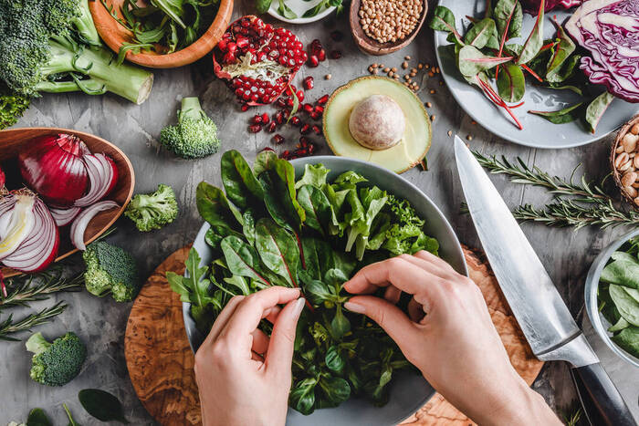 woman preparing a salad with greens and different vegetables scattered around on the table