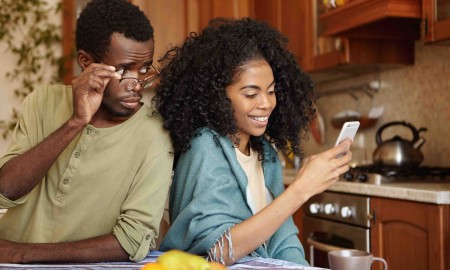 Jealous curious black man holding glasses spying his girlfriend's mobile phone while she is typing message to her lover and smiling happily. Betrayal, unfaithfulness, infidelity and lack of trust