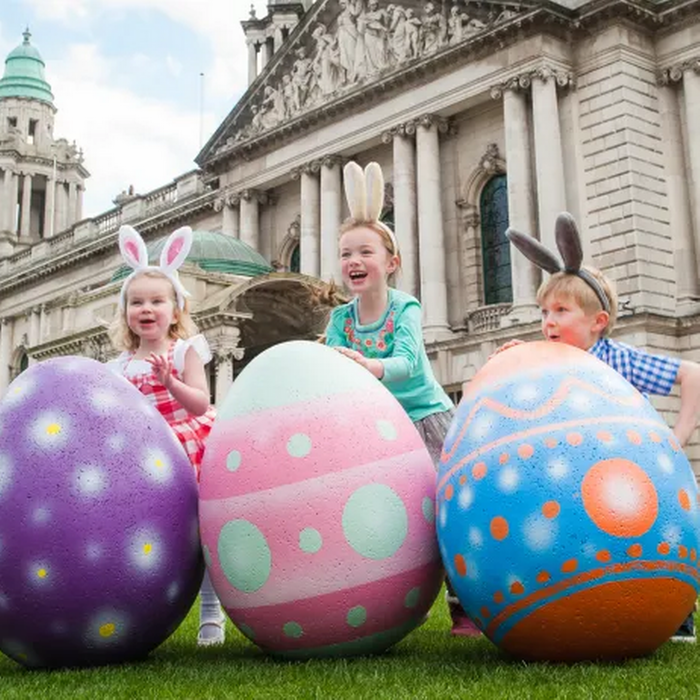 three little girl playing with three large painted eggs on a lawn in front of beautiful buildings