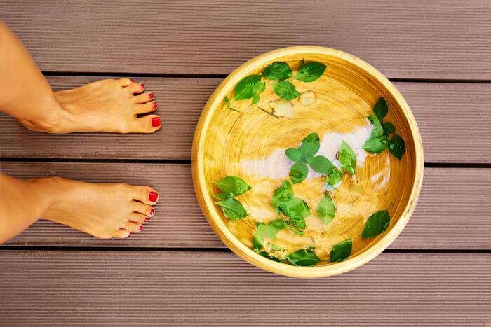 feet with red nail polish standing on a wooden floor in front of a large wooden bowl with herbs floating inside