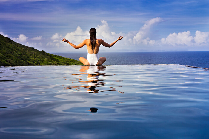 infinity pool woman in white swim suit meditating at the edge with the sea in the background
