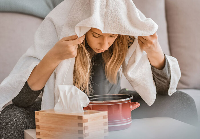 woman with blond hair in gray clothes sitting on a couch with a white towel on her head over a steaming pot inhaling the steam