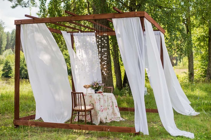 white outdoor curtains on a wooden frame with a table and two chairs in the middle on a green lawn