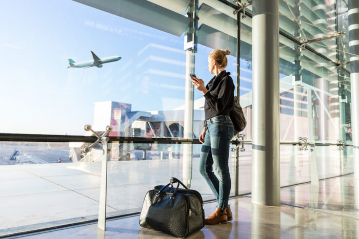 post covid travel blond woman at the airport with her luggage and phone looking from the window at a plane flying