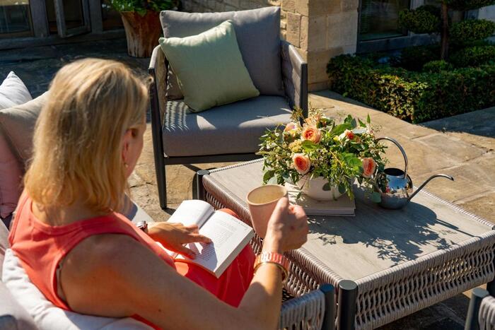 blond woman sitting outside with a book and a cup of tea in a spot with garden furniture and flowers