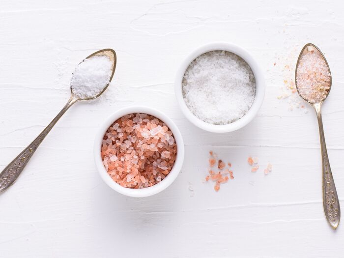 types of salt white and pink salt in white bowls on a white table with two spoons full of salt next to it