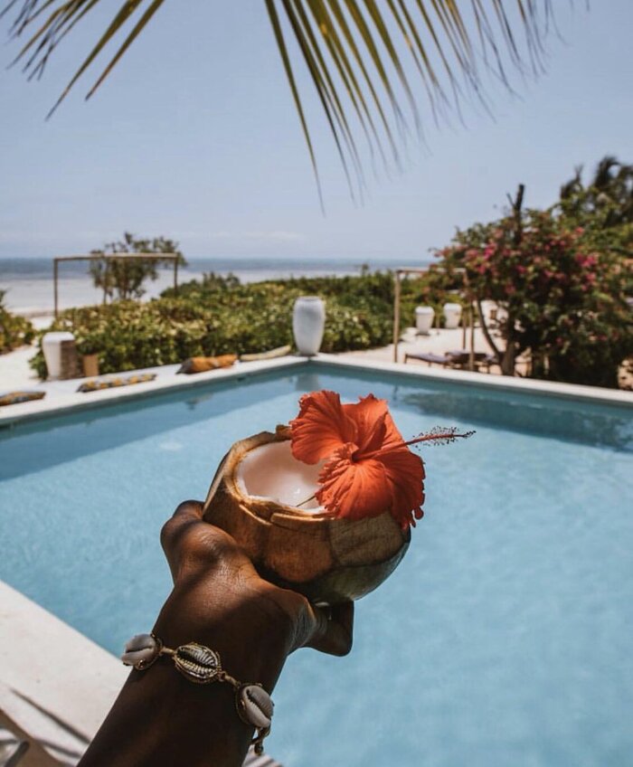 woman's hand holding a small coconut cut in half with an exotic orange flower in it overlooking a pool