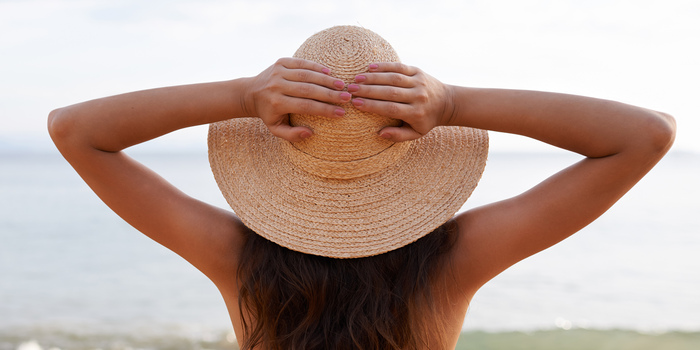 woman with her back to the camera holding her sun hat on her head with both hands looking at the sea