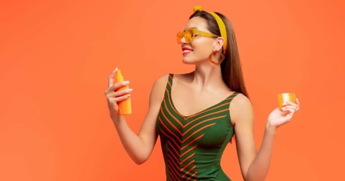 woman in green and orange swimming suit with orange sunglasses and head band on an orange background spraying sun protection lotion on her face and body