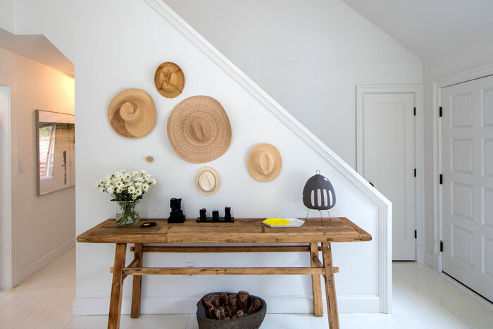 white home entrance furnished with natural materials wooden table and woven hats hanging on the wall