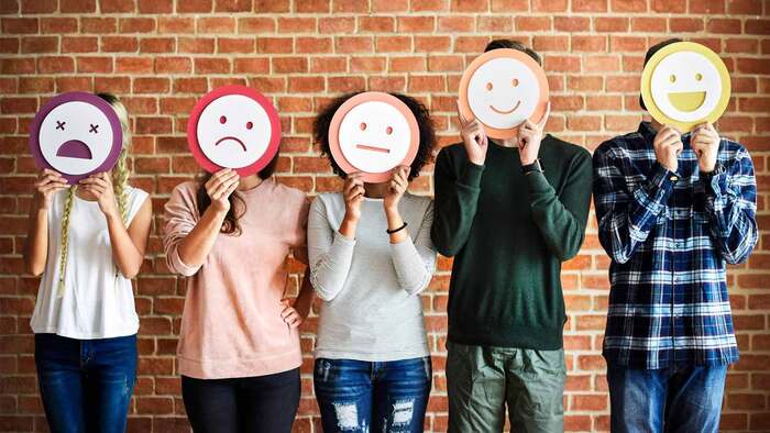 emotional health man and women standing in front of a brick wall holding paper mask emojis with different emotions