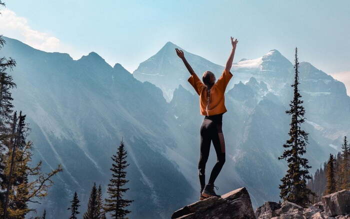 woman in sportswear standing at a cliff on a large rock in the mountains with her hands up in the air