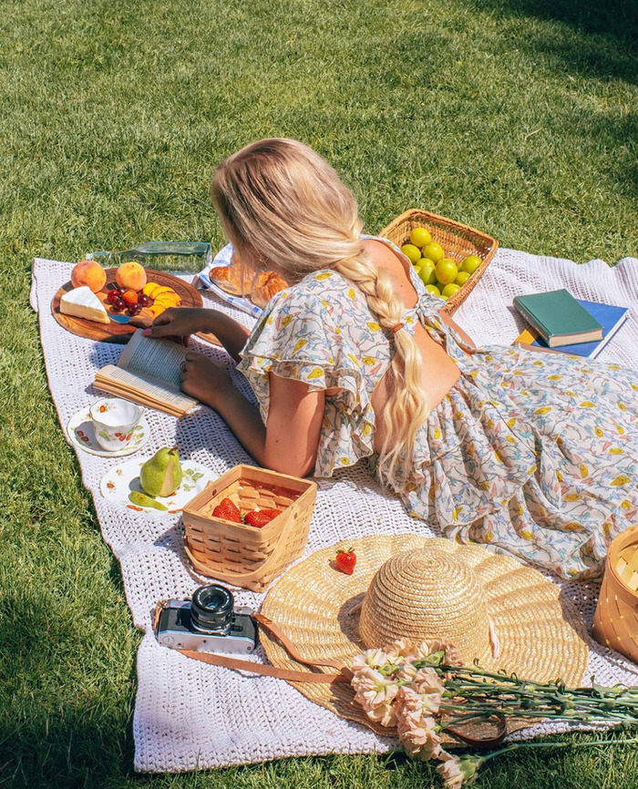 woman in a feminine dress and blond hair lying with her back to the camera on a picnic blanket reading a book and snacking on fruits