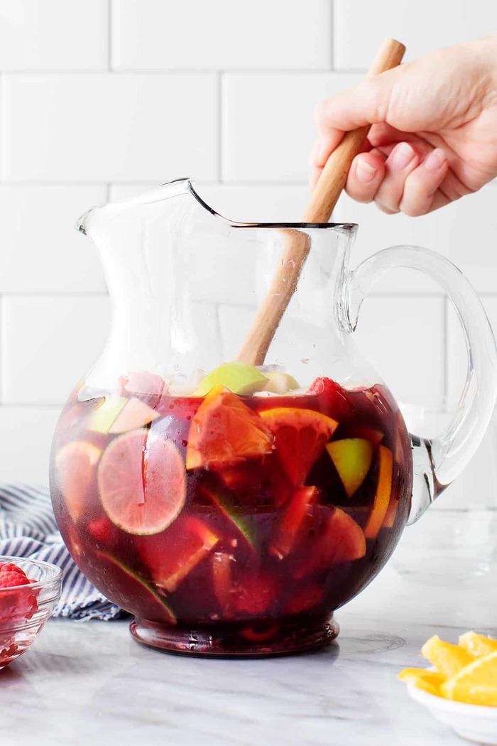 hand stirring a large jug with sangria with a wooden spoon on a white kitchen background