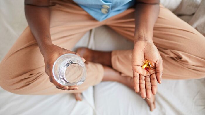 taking supplements woman sitting cross legged on a bed holding a glass of water in one hand and beauty supplements in the other