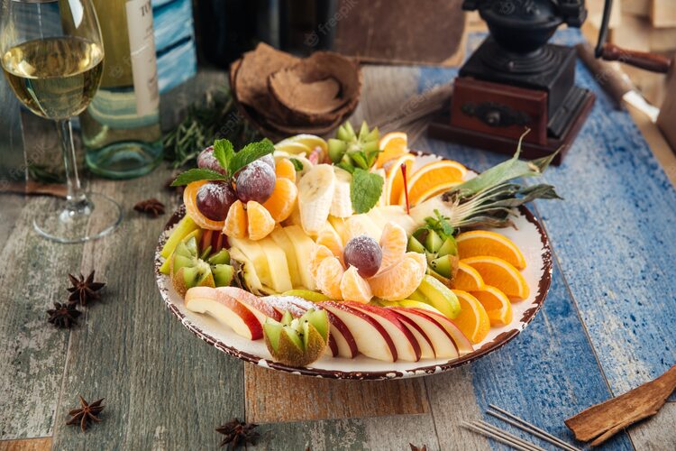 large bowl with fruit salad on a wooden table with spices scattered around a glass of white wine and vintage objects in the background