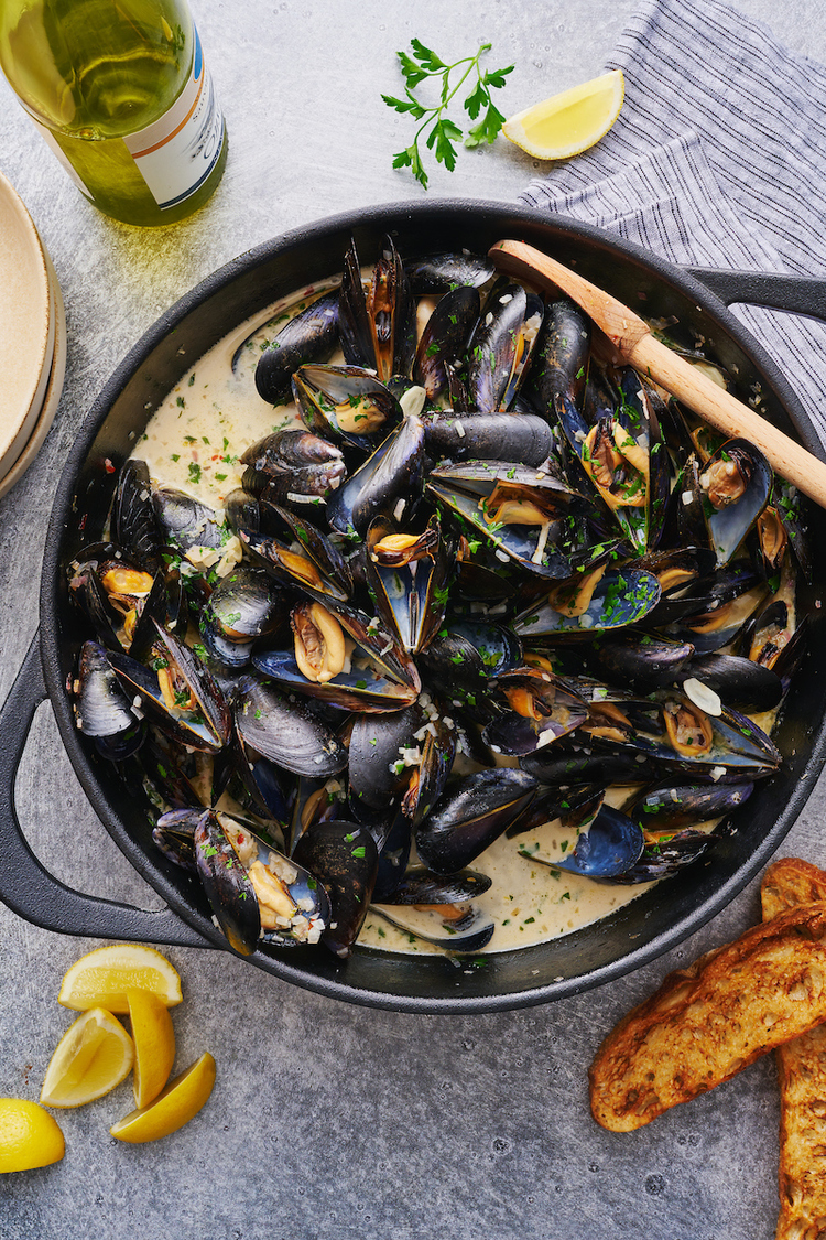 mussels in a large black pan with sauce and a wooden spoon lemons scattered around and a bottle of wine