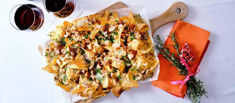 nachos in a large plate on a wooden cutting board with two glasses of red wine decorated with rosemary branches and pomegranate