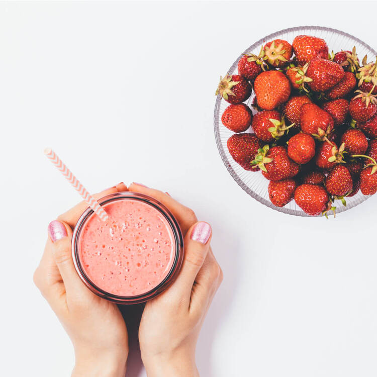 woman holding a glass full of strawberry smoothie with a straw and a glass bowl with fresh strawberries