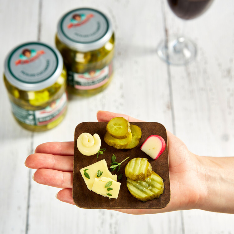pickles and wine woman holding a small wooden square dish with different pickles on it white wooden table surface in the background and two jars of pickes