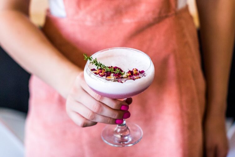 woman in pink dress holding a glass with a rose cocktail with white foam and dried petals on top