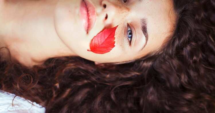 woman with dark curly hair and blue eyes lying back with a red leaf on her cheek