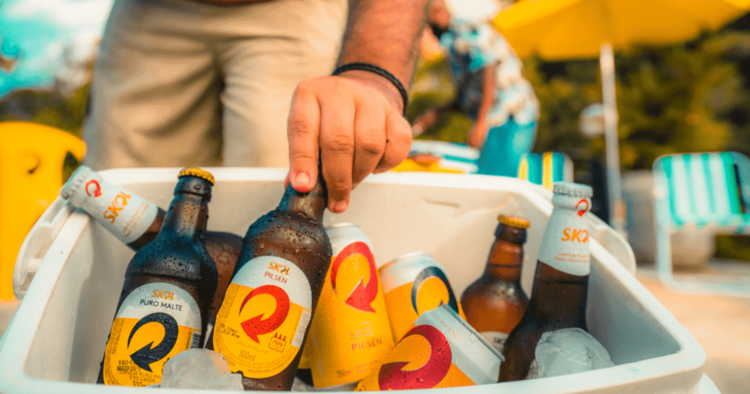 skol beer a man in beige pants picking a bottle of beer from a large plastic cooling bag