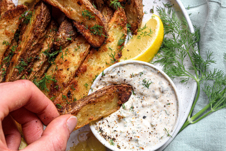 a hand dipping potato wedges in a white yoghurt sauce 